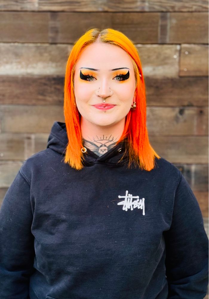 Emma – Portland Barber at Take Pride Barbershop Emma, Portland barber at Take Pride Barbershop with bright orange hair and black hoodie, standing against wood wall